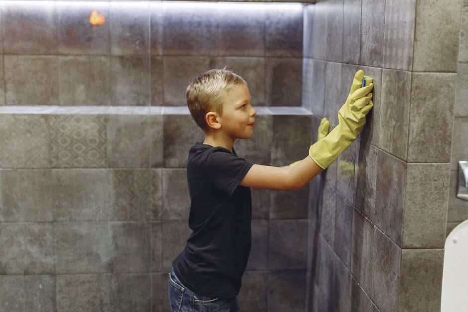 Side view of kid with blond hair in yellow rubber gloves cleaning tile wall in bathroom